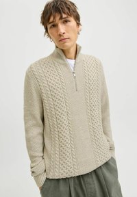 Pullover en tricot de couleur beige clair avec un col zippé à mi-hauteur. Présente un design texturé avec des motifs en torsades et des poignets et un ourlet côtelés.