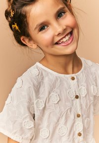 Witte geborduurde blouse met bloemmotieven, korte mouwen en houten knopen, met een zachte textuur en delicate ontwerpelementen.