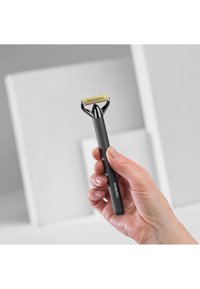 Svart håndtak med et sølvfarget blad og gullakcenter, med en flat egg og små tenner, holdt i en hånd mot en nøytral bakgrunn.