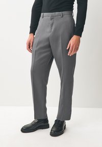 Pantalon gris avec une coupe ajustée, fabriqué en tissu lisse. Il présente des plis à l'avant, des passants pour ceinture et des poches latérales. Associé à des mocassins noirs.