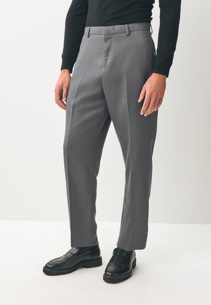 Pantalon gris avec une coupe ajustée, fabriqué en tissu lisse. Il présente des plis à l'avant, des passants pour ceinture et des poches latérales. Associé à des mocassins noirs.