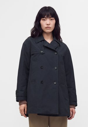 Femme aux longs cheveux foncés porte un manteau noir à double boutonnage avec des boutons par-dessus un pull marron et un pantalon beige sur un fond uni.