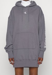 Grå överdimensionerad hoodie med framficka, dragsko och vitt logo på bröstet. Text på ärmen. Bekväm blandning av bomull, avslappnad design.