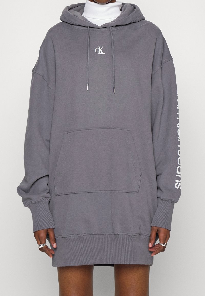 Grå överdimensionerad hoodie med framficka, dragsko och vitt logo på bröstet. Text på ärmen. Bekväm blandning av bomull, avslappnad design.