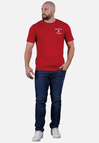 Homme avec une barbe portant un T-shirt rouge Ruckfield, un jean bleu, des baskets blanches, se tenant avec une main dans la poche, regardant à gauche.