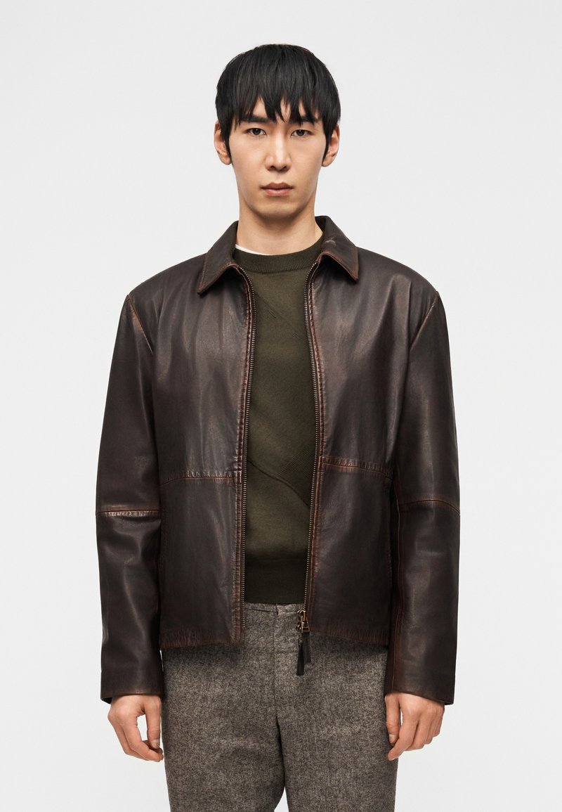 Νεαρός άνδρας που φοράει καφέ σκούρο δερμάτινο μπουφάν πάνω από ένα χακί πράσινο πουλόβερ και γκρι μάλλινες παντελόνες, στέκεται μπροστά σε μονόχρωμο φόντο.