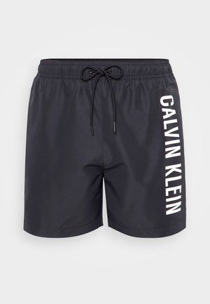 Svart badeshorts med elastisk linning og snøring, med en hvit vertikal "CALVIN KLEIN"-logo på siden. Glatt tekstur.