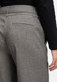 Persoon met grijze visgraatbroek met een slanke achterzak en een zwarte lange mouwtop, hand ontspannen naast zich.
