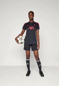 Μαύρη ποδοσφαιρική στολή με κόκκινες λεπτομέρειες, με κοντά μανίκια, ταιριαστές βερμούδες και μαύρα παπούτσια ποδοσφαίρου. Παίκτης κρατάει μια μπάλα ποδοσφαίρου.