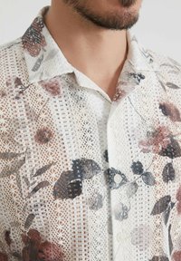 Chemise blanche à manches courtes avec une texture en dentelle et des motifs floraux dans des tons de rose pâle, marron et gris. Elle dispose d'un col standard et de boutons.