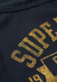 Tissu en coton marine avec un relief vertical présentant un texte imprimé en doré "SUPER" et un design géométrique, mettant en avant la texture et le contraste.