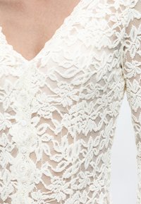 Κοντινό πλάνο ενός λευκού μακρυμάνικου τοπ από δαντέλα με ανθικά σχέδια και μικρά κουμπιά κατά μήκος της μπροστινής πλευράς, που δείχνει ντεκολτέ σε σχήμα V.