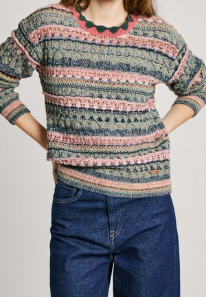 Kvinde iført en flerfarvet strikket sweater med pink, grønne og blå striber, kombineret med højtaljede mørkeblå jeans, hænderne på hoften.