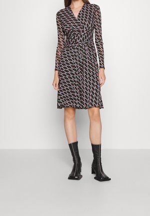 Kobieta w wzorzystej sukience kopertowej do kolan z długim rękawem oraz czarnych botkach na kwadratowym nosku, stojąca na jednolitym tle.