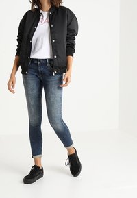 Svart bomberjacka med tryckknappar, vit t-shirt med röd text, blå slim-fit jeans och svarta sneakers, som visar en avslappnad streetwear-stil.