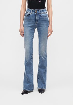 Γυναίκα φορούν ψηλόμεσα, ανοιχτό μπλε τζιν bootcut με δύο καφέ κουμπιά, συνδυασμένα με μαύρα μυτερά παπούτσια και μαύρο τοπ.