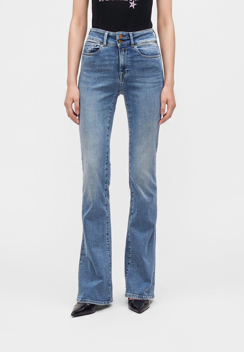 Γυναίκα φορούν ψηλόμεσα, ανοιχτό μπλε τζιν bootcut με δύο καφέ κουμπιά, συνδυασμένα με μαύρα μυτερά παπούτσια και μαύρο τοπ.