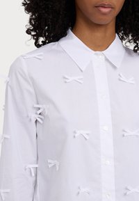 White shirt with a classic collar features multiple small white fabric bows attached to the front and sleeves, made from smooth cotton material.