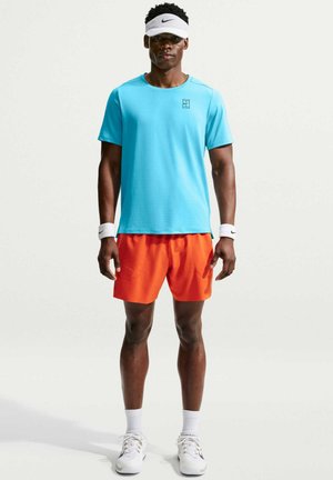 Mann trägt blaues Kurzarmhemd, leuchtend orangefarbene Shorts, weiße Sportschuhe, Armbänder und ein weißes Nike-Visier und steht vor einem einfarbigen Hintergrund.