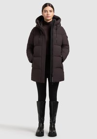 Zwarte pufferjack met een capuchon, voorzien van een rits aan de voorkant, meerdere zakken en een gegoten ontwerp. Gedragen over een donkere turtleneck en leggings.