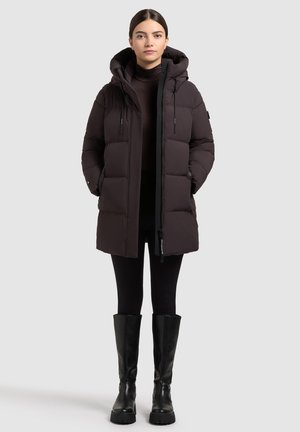 Zwarte pufferjack met een capuchon, voorzien van een rits aan de voorkant, meerdere zakken en een gegoten ontwerp. Gedragen over een donkere turtleneck en leggings.