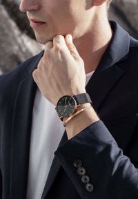 Czarny zegarek z siatkowym paskiem i różowozłotymi akcentami na nadgarstku, w połączeniu z cienką metalową bransoletką. Granatowa marynarka z widocznymi guzikami.