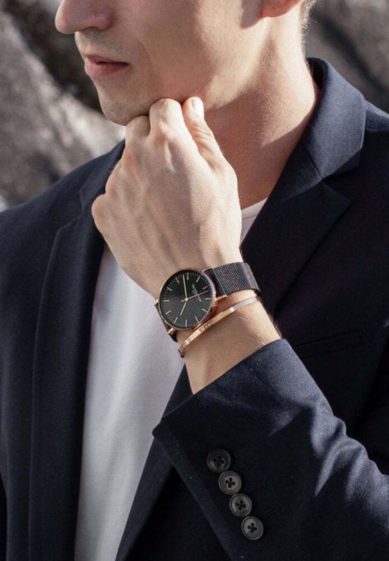 Czarny zegarek z siatkowym paskiem i różowozłotymi akcentami na nadgarstku, w połączeniu z cienką metalową bransoletką. Granatowa marynarka z widocznymi guzikami.