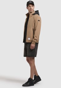 Chaqueta de caparazón blando de color marrón claro con capucha negra, bolsillos con cremallera y puños elásticos, combinada con pantalones cortos negros y zapatillas altas.
