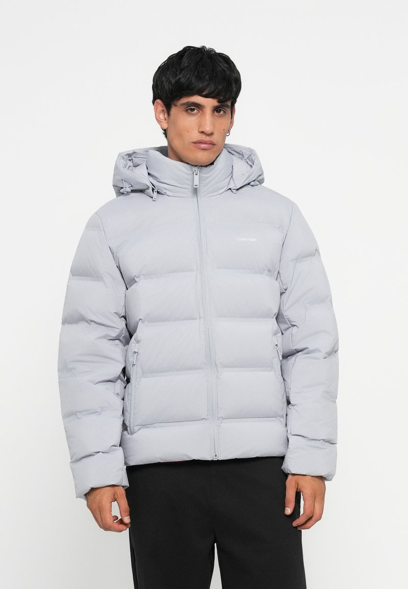 Light gray puffer jacket with a zipper and adjustable hood. Features quilted sections and a simple logo on the chest. Smooth texture.