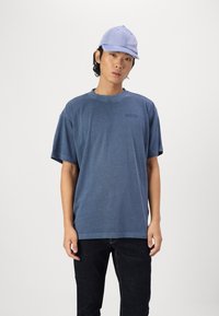 Blå kortermet T-skjorte med avslappet passform og rund hals, med en subtil logo på brystet. Brukt med mørke jeans og en lyseblå caps.