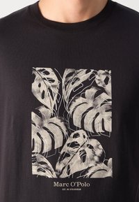 T-shirt nera con stampa color crema di foglie tropicali, con dettagli fini e testo in basso che recita "Marc O'Polo."
