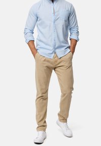Hellblaues, langärmeliges Hemd mit Knöpfen und einer Tasche, kombiniert mit beigen Hosen und weißen Sneakern. Glatter Stoff, lockere Passform.