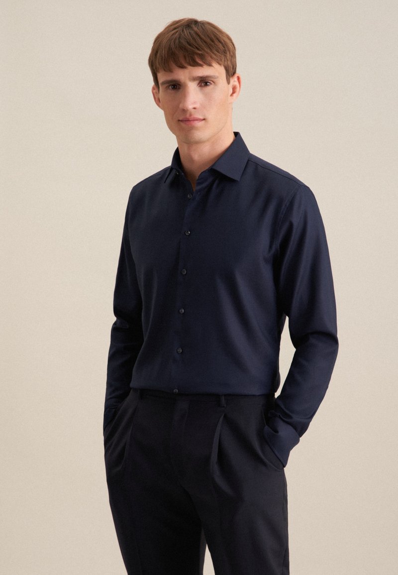 Camicia a maniche lunghe blu navy in tessuto morbido, con colletto classico e patta con bottoni, indossata con pantaloni eleganti scuri.
