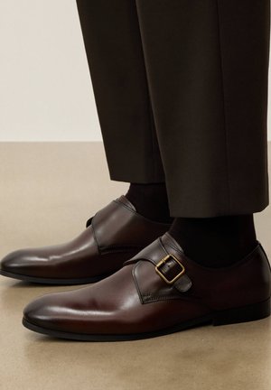 Čevlji za moške iz temno rjavega usnja z monaško sponko v zlati barvi, nošeni s črnimi nogavicami in temnimi hlačami na bež tleh.
