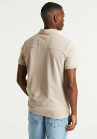 Polo beige con una morbida consistenza, maniche corte e colletto. Presenta un design con yoke posteriore e un orlo dritto. Abbinato a jeans azzurri chiari.