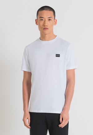 Camiseta de algodón blanca con escote redondo, mangas cortas y un parche de logo rectangular negro en el lado izquierdo del pecho. Diseño simple, sin patrones.