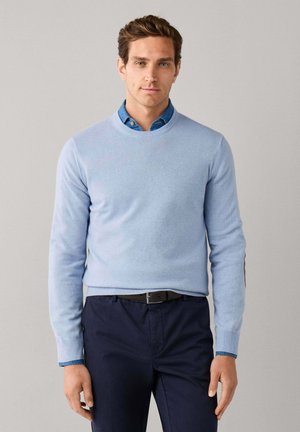 Homme portant un pull bleu clair par-dessus une chemise bleue à col, avec un pantalon bleu marine et une ceinture noire, debout devant un fond gris uni.