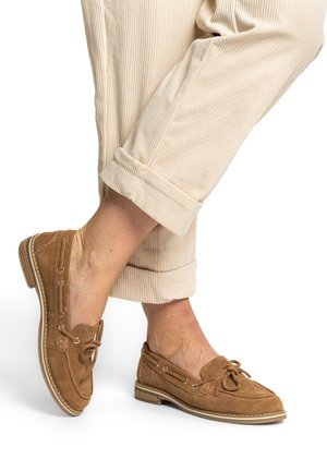 Feet wearing tan suede lace-up loafers paired with beige corduroy pants rolled at the cuffs, shown against a white background.