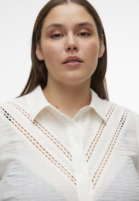 Blusa blanca con cuello, que presenta detalles de corte en los hombros y el pecho. La tela es ligera y ligeramente texturizada.