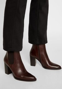Bottines en cuir marron à bout pointu, avec des panneaux latéraux élastiques et un talon carré haut, associées à un pantalon noir ajusté.