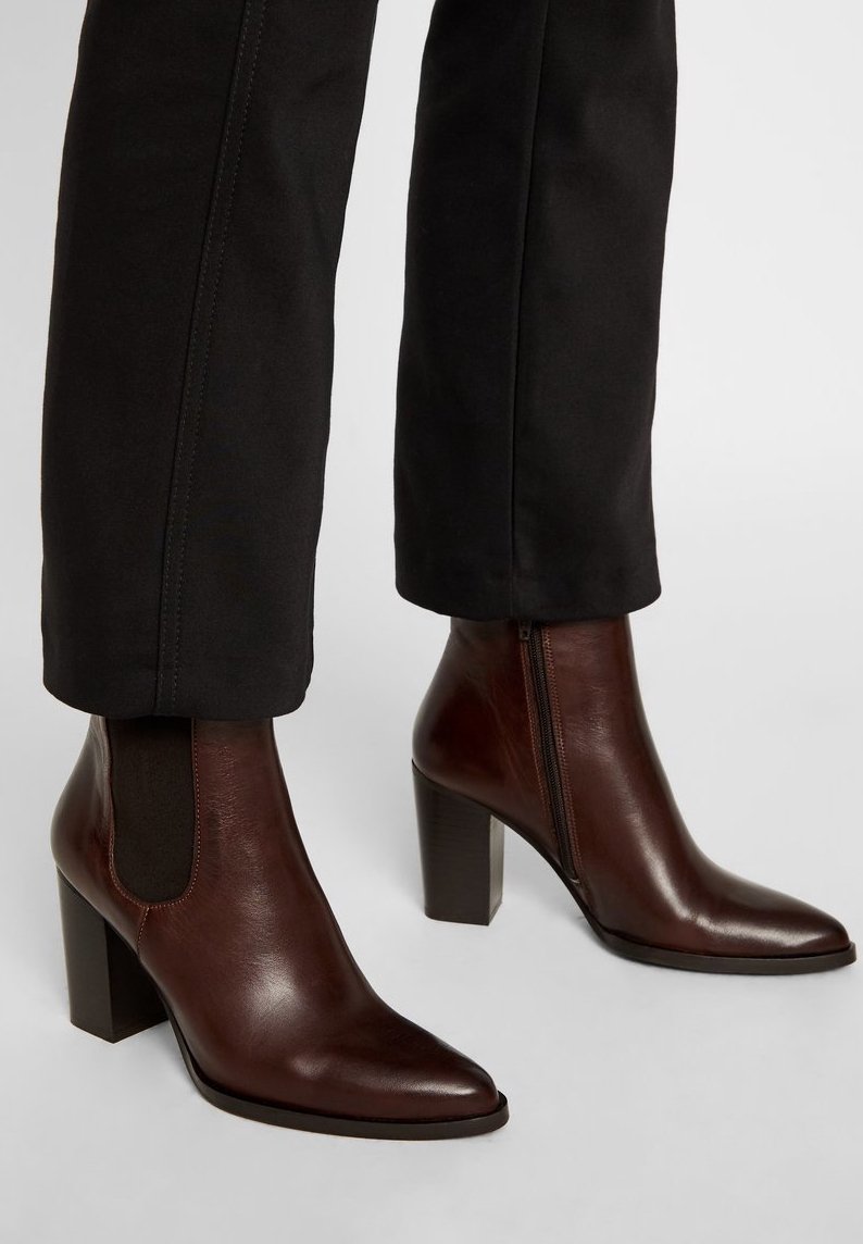 Bottines en cuir marron à bout pointu, avec des panneaux latéraux élastiques et un talon carré haut, associées à un pantalon noir ajusté.