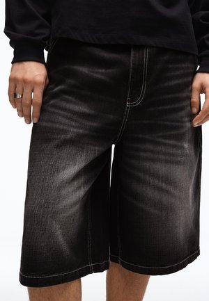 Person trägt schwarze, gewaschene Denim-Shorts in Kniehöhe mit kontrastierenden weißen Nähten und ein schwarzes Langarmshirt, die Hände liegen seitlich am Körper.