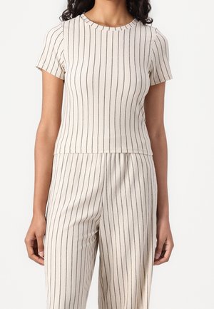 Nainen, jolla on päällään beige lyhythihainen paita ja siihen sopivat housut, joissa on ohuet pystysuorat mustat raidat valkoisella taustalla.