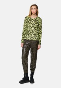 Langärmliges Oberteil im grün-schwarzen Leopardenmuster, kombiniert mit dunkler, glänzender Cargo-Hose mit elastischen Bündchen und seitlichen Taschen. Schwarze Stiefel.