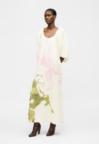 Γυναίκα με κοντά μαλλιά που φοράει ένα φαρδύ, μακρύ φόρεμα σε κρέμ απόχρωση με ροζ και πράσινα αφηρημένα σχέδια αλόγων, που στέκεται με τα χέρια πίσω από την πλάτη και καφέ ψηλοτάκουνα.