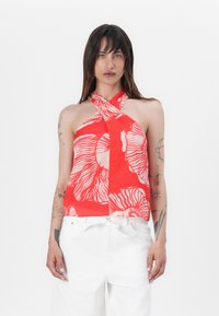 Red halter top with a floral pattern in white, featuring a crossover design at the neck. Worn with white high-waisted pants.