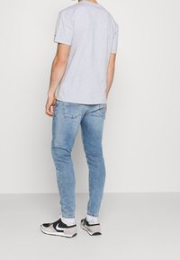 Mies, který má na sobě světle šedé tričko, světle modré slim fit džíny, bílé ponožky a černo-bílé tenisky Nike, stojí proti jednobarevnému pozadí.