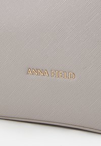 Borsa a mano grigia chiara con una finitura texturizzata. Presenta il marchio "ANNA FIELD" in oro impresso sulla superficie.