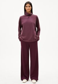 Langer lila Pullover mit hohem Kragen und bestickten Details, kombiniert mit weit geschnittenen passenden Hosen, ergänzt mit cremefarbenen Schuhen.
