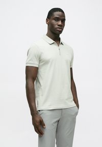 Gaiši pelēka polo krekls ar rāvējslēdzēja apkakli, īsām piedurknēm un nelielu logotipu uz krūtīm, kopā ar gaiši pelēkām biksēm. Gluda auduma faktūra.